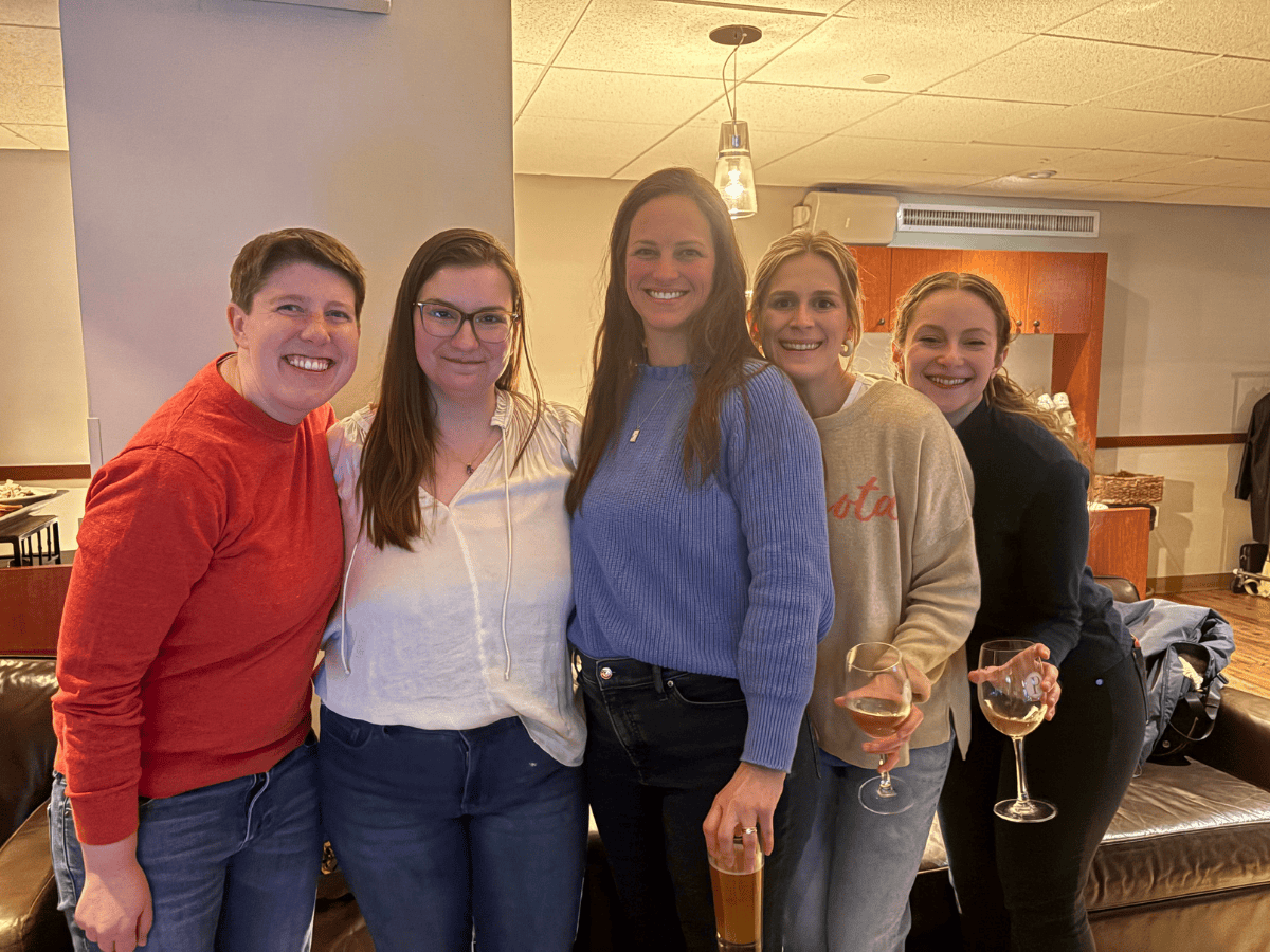 Five people in casual clothing smiling and holding beverages in a well-lit bowling alley.