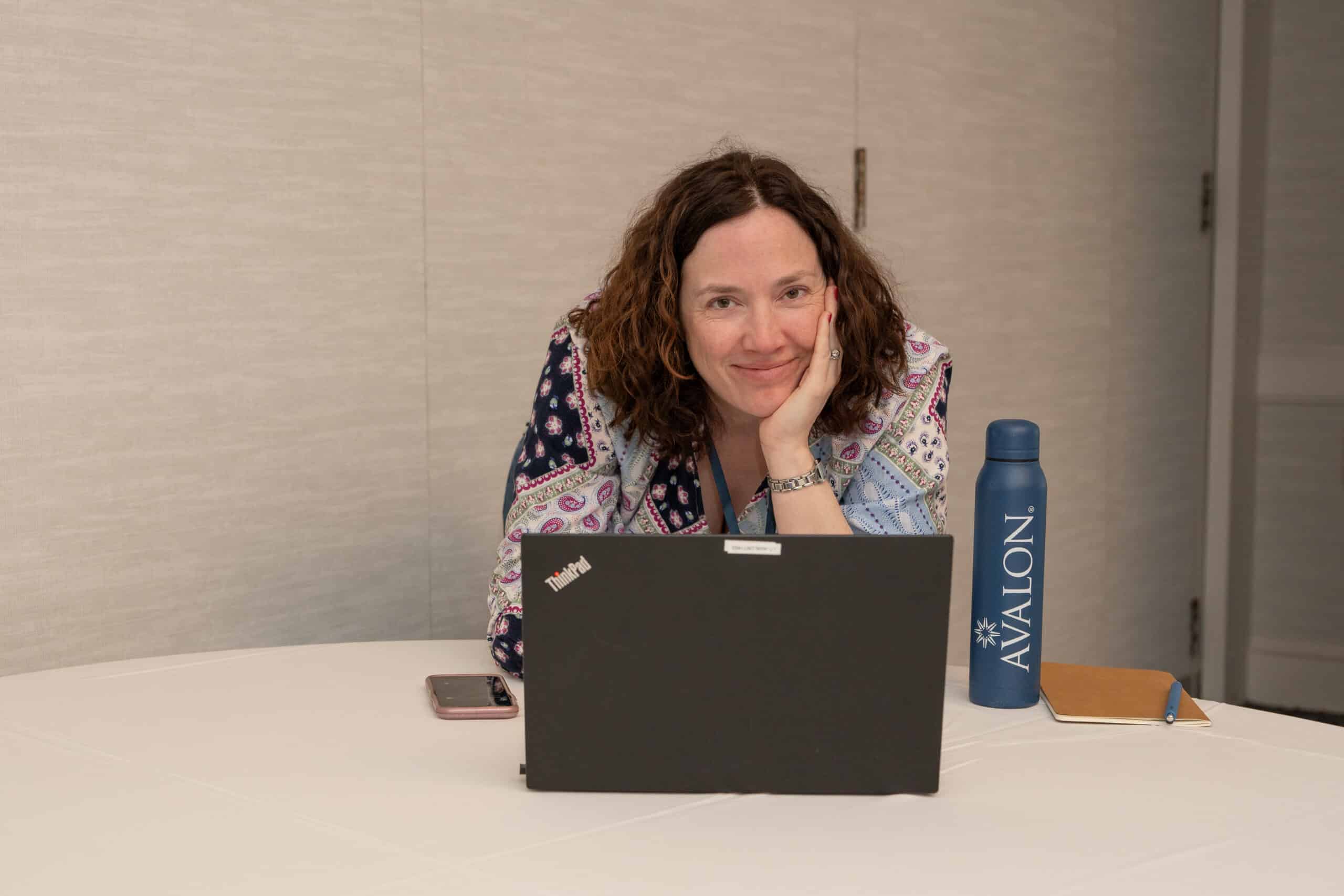 A woman leaning over a table and working on her laptop. Her head is in her left hand and she's looking at the camera and smiling. Next to her is an Avalon-branded navy blue water bottle.