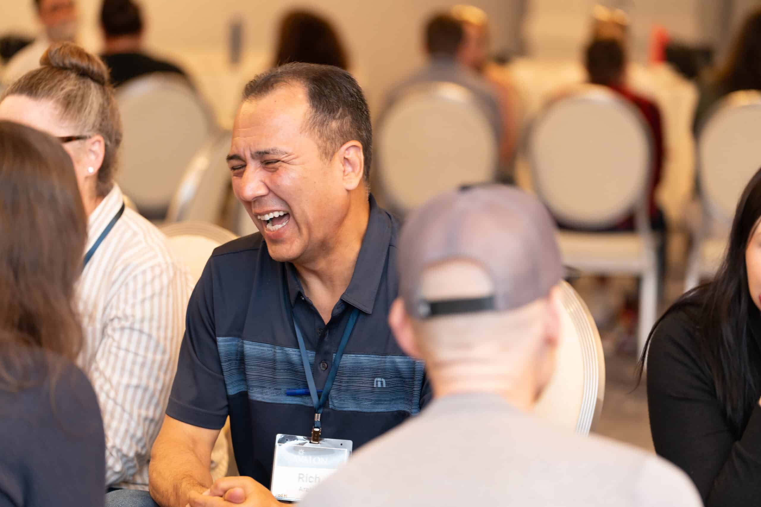 A man wearing a navy blue shirt and name badge is laughing while sitting at a table with others in a digital conference or meeting room. Several people are seated and engaged in conversation around him.