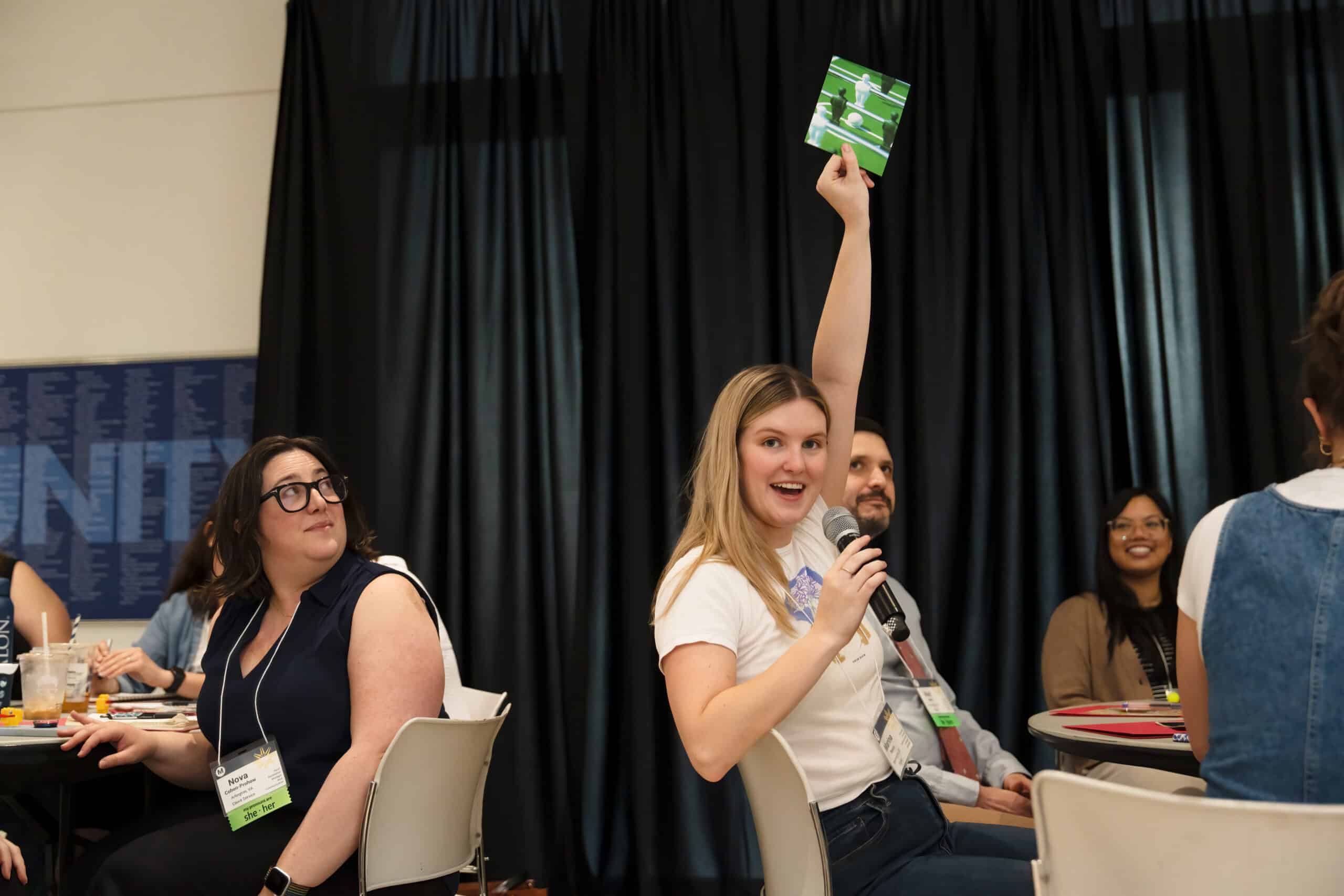 A woman at a table raises her hand holding a green card while speaking into a microphone, possibly discussing direct mail strategies. Several others are seated around her, all facing the front of the room.