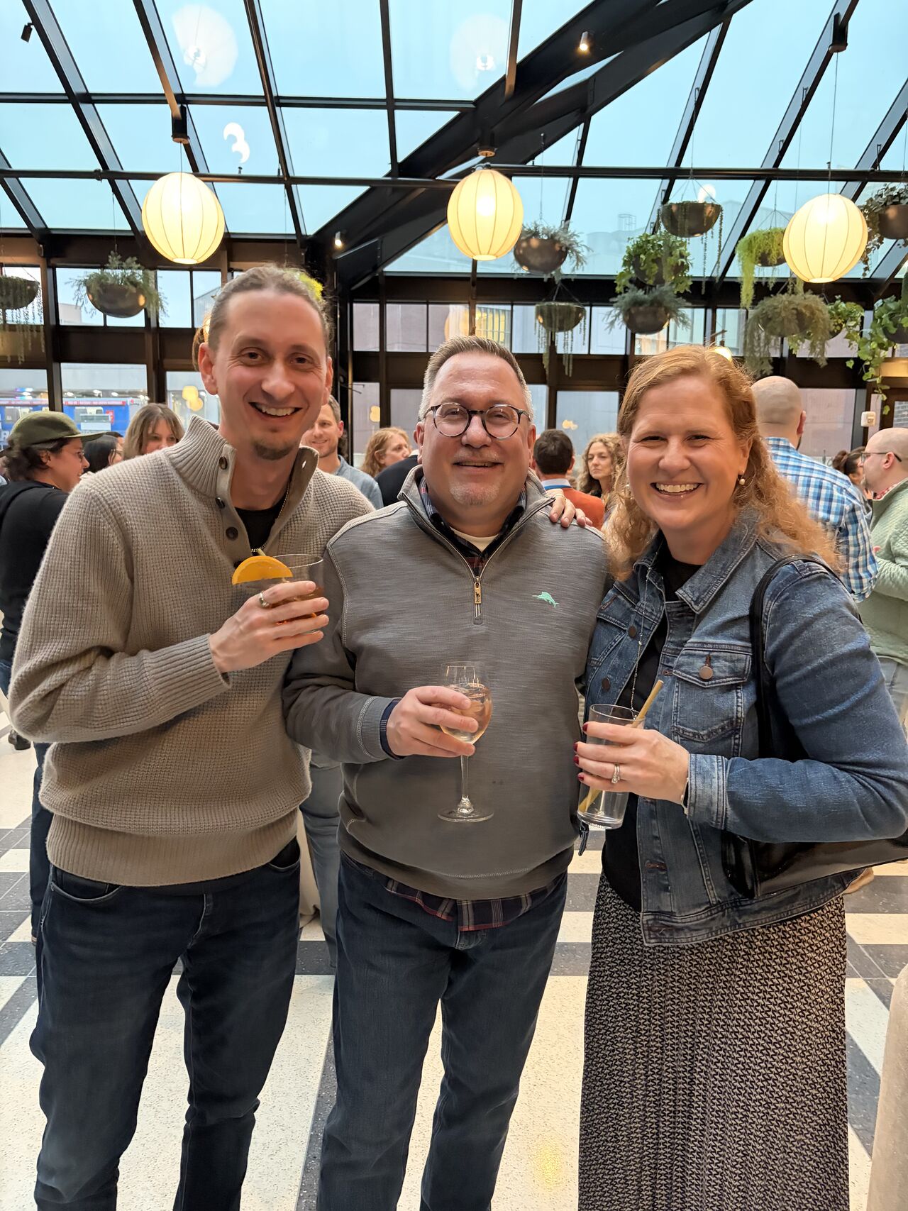 David, John, and Kerri standing indoors at a social gathering, each holding a drink.