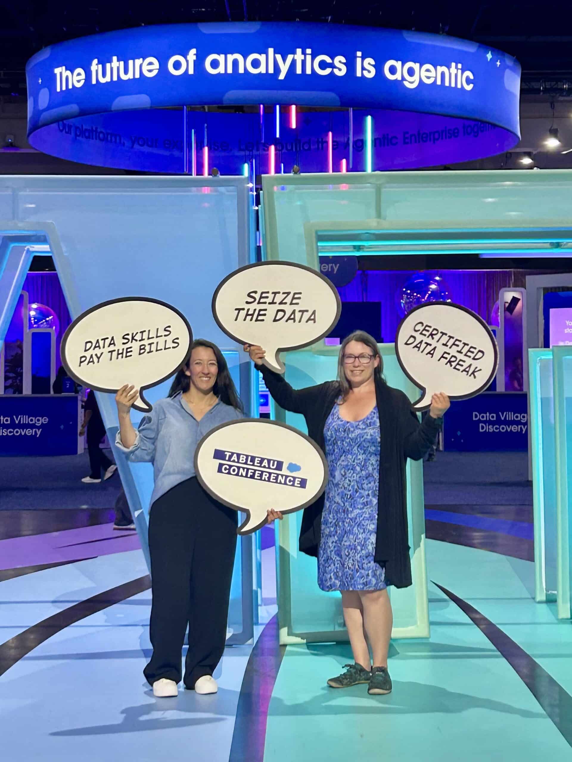 Photograph of two people at a Tableau Conference event holding speech bubble signs with phrases "DATA SKILLS PAY THE BILLS," "SEIZE THE DATA," "CERTIFIED DATA FREAK," and "TABLEAU CONFERENCE." Background features large illuminated text reading "The future of analytics is agentic" and colorful exhibition booth structures.