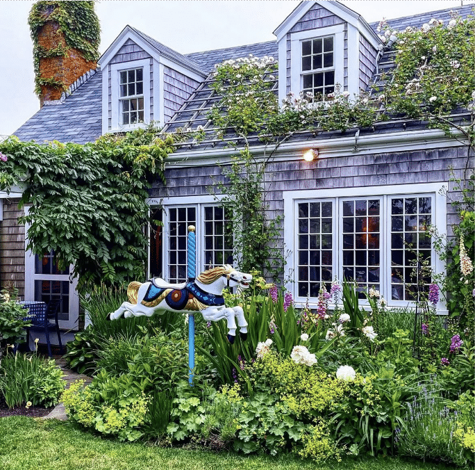 Photograph of a charming cottage with gray shingle siding, white-framed windows, and a roof with two dormer windows, surrounded by lush greenery and flowering plants. A decorative carousel horse mounted on a blue pole stands prominently in a vibrant garden filled with various flowers and shrubs in front of the house.