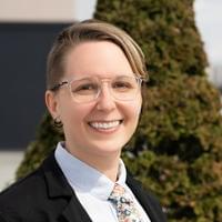 Sarah has short blonde hair, clear-rimmed glasses and two black hoops in her ear. Sarah is wearing a black blazer, white shirt, and flowered tie.