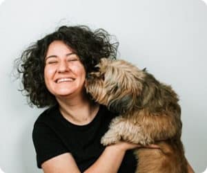 Person with dark curly hair wearing a black shirt and holding a small brown dog that is sniffing their face.