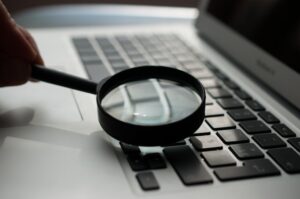 A hand holds a magnifying glass over the keyboard of a laptop, focusing on several keys. The laptop is silver with black keys, and the image is taken in close-up.