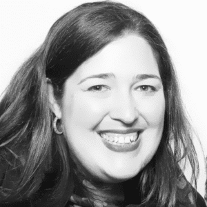 A woman with shoulder-length dark hair is smiling at the camera. She is wearing hoop earrings and a dark top. The photo is in black and white with a plain background.