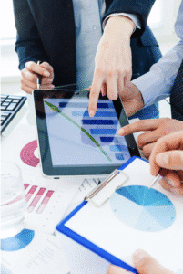 Three people in business attire point at a tablet displaying a bar graph with a downward trend, surrounded by documents and charts on a table. One person holds a clipboard with a pie chart, highlighting their analysis of recent data.