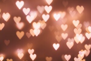 A blurred image of multiple heart-shaped lights in various sizes on a soft, warm brown background. The lights are out of focus, creating a bokeh effect.