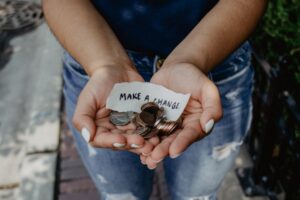 A person holds coins and a small piece of paper with Make a Change! written on it in their open hands. The individual is wearing blue jeans and has light-colored nail polish.