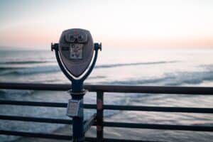 A coin-operated binocular viewer is mounted on a pier railing, facing the ocean with gentle waves and a pastel-colored sky at either sunrise or sunset.