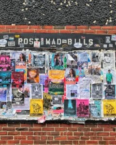A brick wall covered with colorful posters for concerts and events, layered over each other beneath a weathered black sign that reads POST NO BILLS with some letters partially missing.